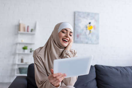 excited muslim woman laughing near digital tablet at home