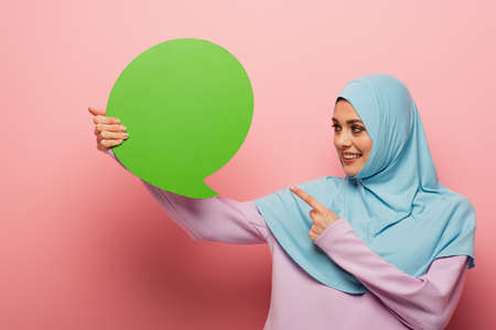 smiling muslim woman pointing at green thought bubble on pink background