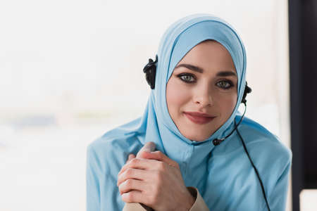 pretty muslim woman in hijab and headset smiling at camera in officeの写真素材