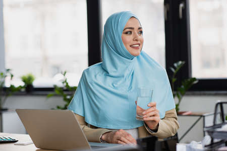 successful muslim businesswoman with glass of water looking away near laptop in officeの写真素材
