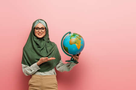 smiling muslim teacher in green hijab pointing at globe on pink backgroundの写真素材