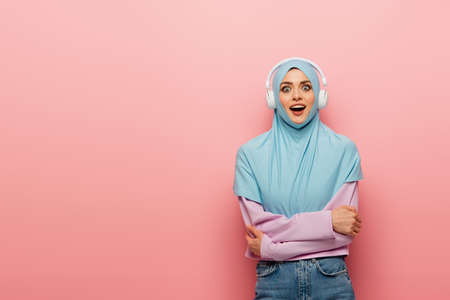 amazed arabian woman in blue hijab and headphones looking at camera on pink backgroundの写真素材