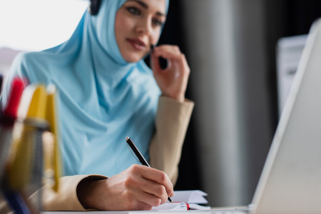 blurred muslim woman writing in notebook while working in call centerの写真素材