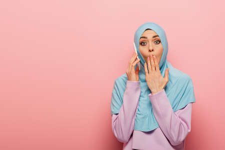 shocked arabian woman in blue hijab talking on cellphone on pink backgroundの写真素材