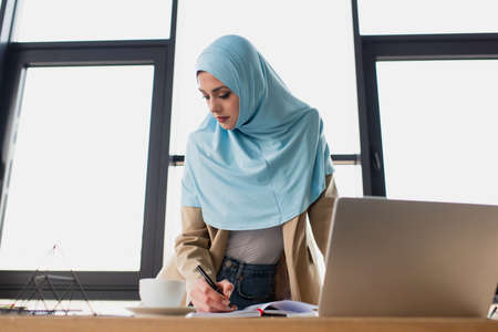 pretty muslim businesswoman writing in notebook while standing near laptopの写真素材