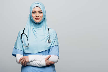 joyful muslim doctor in blue hijab looking at camera while standing with crossed arms isolated on grayの写真素材