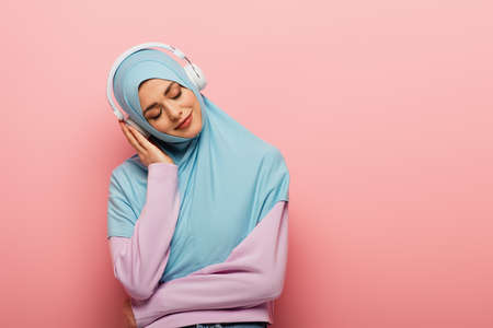 pleased muslim woman listening music in headphones with closed eyes on pink background