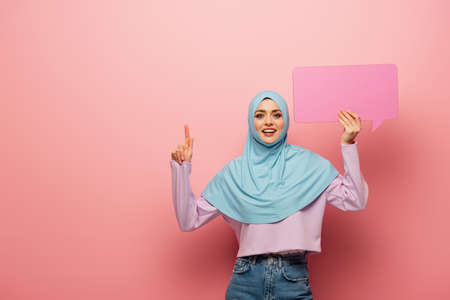 positive muslim woman pointing up with finger while holding speech bubble on pink backgroundの写真素材