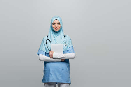 young arabian doctor in blue hijab and uniform holding digital tablet isolated on grayの写真素材