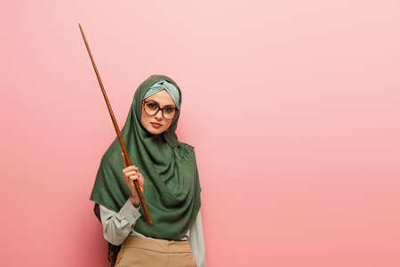 young arabian teacher in green hijab holding pointing stick isolated on pinkの写真素材