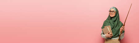 happy muslim teacher with pointing stick and books looking away on pink background, bannerの写真素材