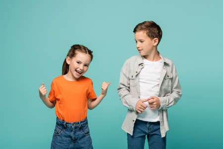 happy girl in orange t-shirt pointing with thumb at boy isolated on blueの写真素材