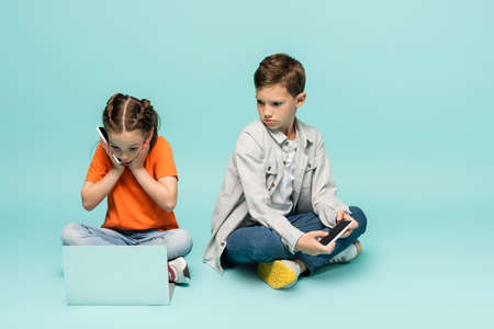 shocked girl talking on smartphone and using laptop near sad boy on blueの写真素材