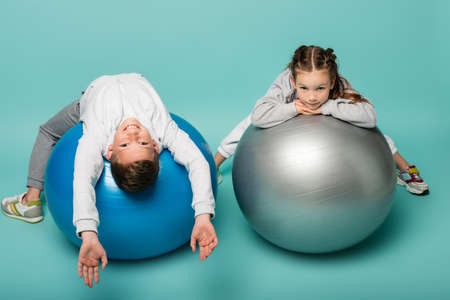 happy boy in sportswear lying on fitness ball near girl on blueの写真素材