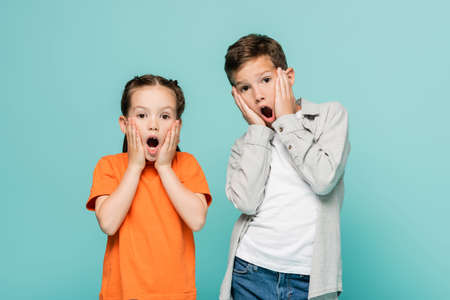 shocked kids touching cheeks and looking at camera isolated on blue