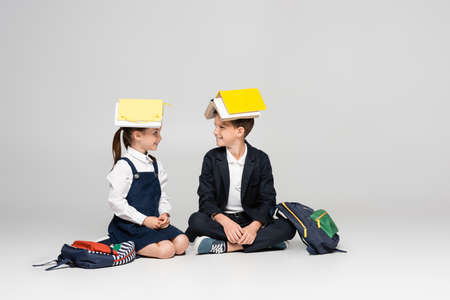 happy schoolkids in uniform with books on heads sitting and looking at each other on gray