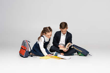 schoolkids in uniform sitting and reading books near backpacks on grayの写真素材
