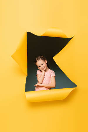 pleased kid looking at camera while smiling through ripped hole on yellow backgroundの写真素材