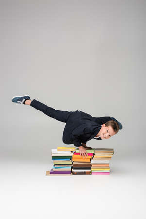 happy schoolboy doing handstand on pile of books on gray