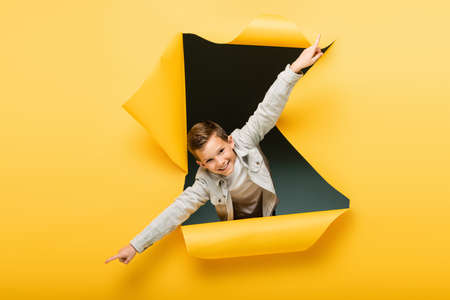 joyful boy with outstretched hands looking at camera through ripped hole on yellow backgroundの写真素材