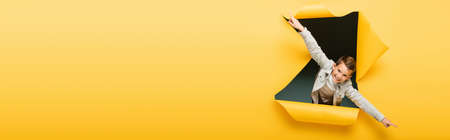 joyful boy with outstretched hands looking at camera through ripped hole on yellow background, bannerの写真素材