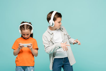 girl using smartphone near boy in wireless headphones listening music while dancing isolated on blueの写真素材