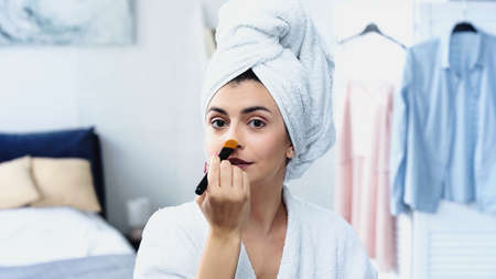 young woman with head wrapped in towel applying face foundation on nose in bedroomの写真素材
