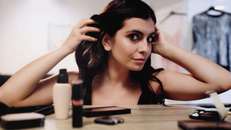 brunette young woman adjusting hair with hands near table with decorative cosmetics in bedroom