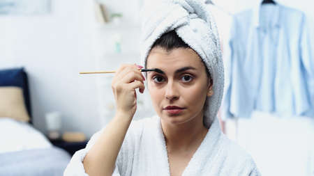 young woman in bathrobe with head wrapped in towel styling eyebrow with brush and looking at camera...