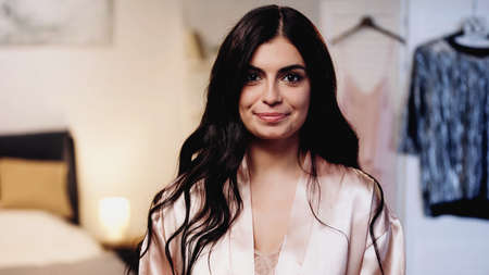smiling young woman in pink silk peignoir with long hair looking at camera in bedroom