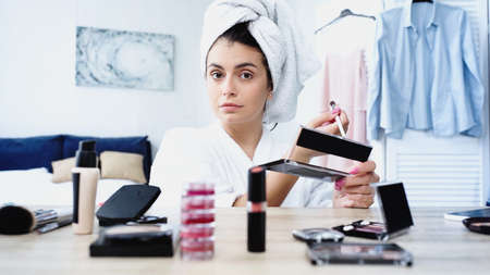 young woman in bathrobe sitting with cosmetic brush and eye shadows near table with decorative cosmetics in bedroomの写真素材
