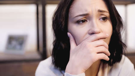 portrait of depressed brunette woman in beige sweater covering mouth with hand at homeの写真素材