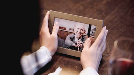partial view of female hands holding photo frame with picture of elderly man at homeの写真素材