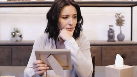 upset woman sitting on table with hand near face and holding photo frame at homeの写真素材