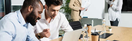 interracial businessmen looking at laptop near businesswomen working during meeting, banner