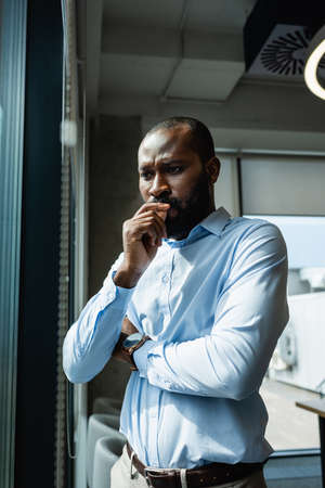 pensive african american businessman holding hand near face while standing in officeの写真素材