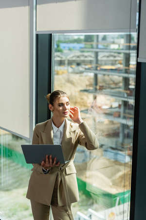 dreamy businesswoman holding laptop while looking away through window in officeの写真素材