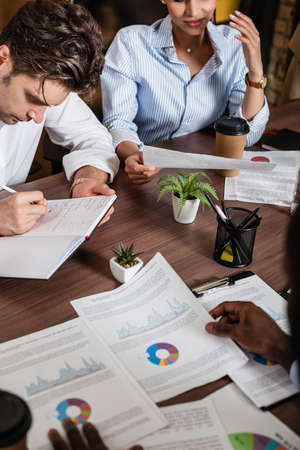 young businessman writing in notebook near papers with analytics and interracial colleaguesの写真素材