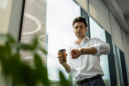 businessman with coffee to go looking at wristwatch near window on blurred foregroundの写真素材