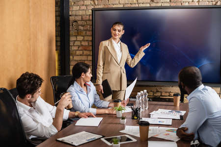 businesswoman pointing at lcd monitor near multiethnic colleagues during meeting