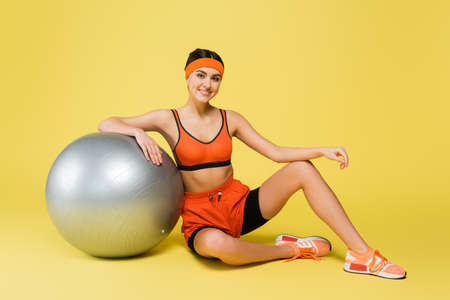 pretty sportive woman looking at camera while sitting near fitness ball on yellow backgroundの写真素材