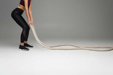 cropped view of sportswoman in black leggings exercising with battle ropes on gray background