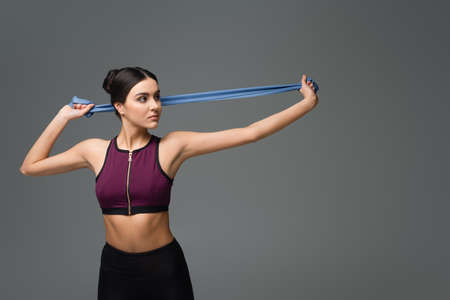 young sportswoman training with resistance band isolated on gray