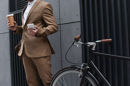 cropped view of businessman holding paper cup and smartphone near bicycleの写真素材