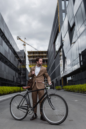 full length of businessman in suit holding bag and standing near bicycle outsideの写真素材