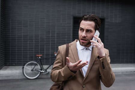 stressed young businessman in formal wear talking on smartphone outside
