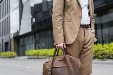 cropped view of businessman in suit holding leather bag and standing with hand in pocket on urban streetの写真素材