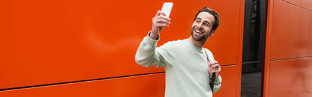 cheerful man in sweatshirt taking selfie on smartphone near orange wall, bannerの写真素材
