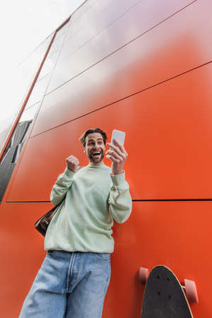 low angle view of amazed man in sweatshirt holding smartphone near orange wall