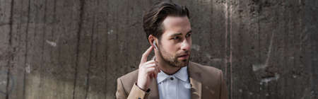 bearded businessman adjusting earphones near building, banner